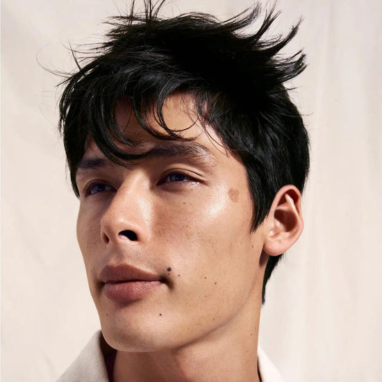 Man with tousled black hair, clean face, and white shirt gazing into the distance with a neutral background.